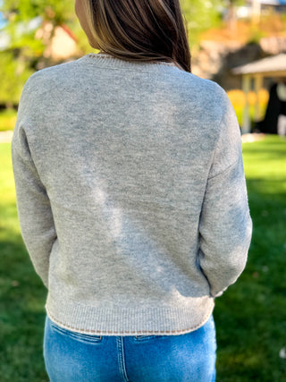 A person viewed from behind wearing a light heather gray knit sweater and blue jeans, standing outdoors on a grassy lawn with a blurred background.