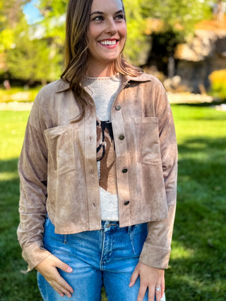 A woman wearing a light brown suede cropped jacket over a graphic t-shirt and blue jeans, standing outdoors on a grassy lawn with trees in the background.