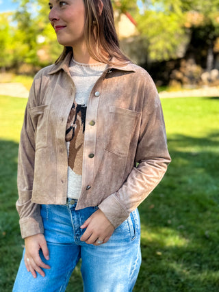 A woman wearing a cropped, light brown suede button-up jacket over a graphic t-shirt and blue jeans, standing outdoors on a grassy lawn.