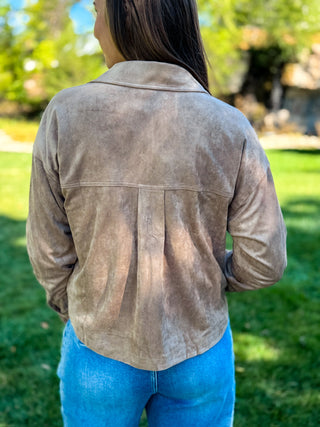 A woman viewed from behind wearing a light brown suede or faux-suede cropped jacket with a pleated back panel, paired with blue jeans, standing outdoors on a grassy lawn.