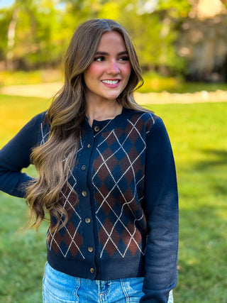 A woman wearing a navy blue button-up cardigan with an argyle pattern in brown, rust, and white, standing outdoors in a grassy park setting.