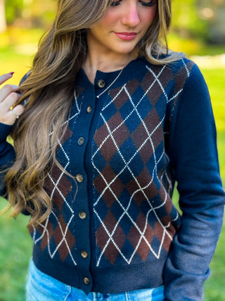 A woman wearing a navy blue cardigan with an argyle pattern in brown, burgundy, and white, photographed outdoors in a grassy setting.