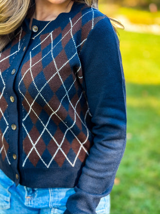 A close-up of a person wearing a navy blue button-front cardigan with an argyle pattern in brown and white, paired with light blue jeans, outdoors on a grassy lawn.