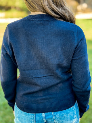A person viewed from behind wearing a dark navy blue ribbed knit sweater and light blue jeans, standing outdoors on a grassy background.