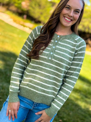 A woman smiling outdoors wearing a sage green and white striped henley sweater with light blue buttons, paired with blue jeans.