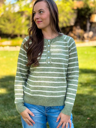 A woman wearing a sage green and white striped knit henley sweater with light blue buttons, paired with blue jeans, standing outdoors on a grassy lawn with trees in the background.