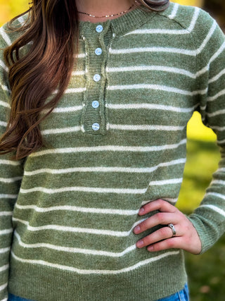 A close-up of a person wearing a knit henley sweater with horizontal olive green and cream stripes, featuring a buttoned placket and ribbed cuffs.