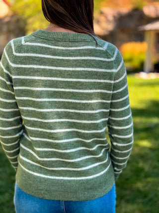 A person viewed from behind wearing a green knit sweater with thin white horizontal stripes, paired with blue jeans, set against a blurred outdoor background of grass and trees.