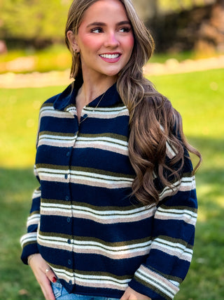 A woman wearing a navy blue button-up cardigan with horizontal stripes in cream, olive green, and tan, standing outdoors in a grassy park setting.