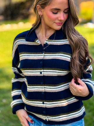 A woman wearing a fuzzy knit button-up cardigan with horizontal stripes in navy blue, beige, and olive green, photographed outdoors in a grassy park setting.