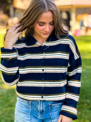 A woman wearing a navy blue knit button-up cardigan with horizontal stripes in olive green, cream, and white, paired with blue jeans, photographed outdoors on a grassy lawn.