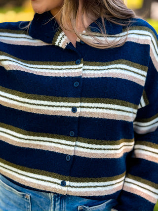 A close-up of a person wearing a navy blue knit button-front cardigan with horizontal stripes in cream, olive green, and light pink, paired with blue jeans.