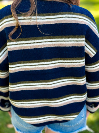 A close-up back view of a person wearing a navy blue knit sweater with horizontal stripes in olive green, cream, and light beige, paired with denim shorts.
