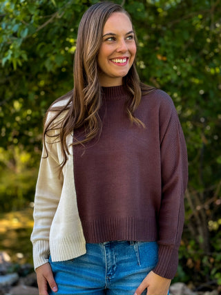 A woman wearing a color-block knit sweater with a dark brown body and cream sleeves, paired with blue jeans, standing outdoors in front of a blurred green foliage background.