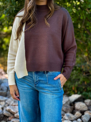 A woman wearing a color-block knit sweater with a brown body and an off-white sleeve, paired with blue jeans, standing outdoors in front of a blurred green background.