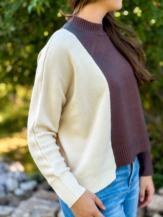 A woman wearing a color-block knit sweater with an off-white left side and sleeve, and a dark plum-brown right side and shoulder, paired with blue jeans in an outdoor setting with green foliage.