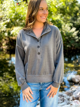 A woman wearing a heather gray long-sleeve quarter-placket henley sweatshirt and blue jeans, standing outdoors in front of a blurred green forest background.