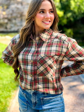 A woman wearing a long-sleeve plaid button-up shirt in beige, red, and black, paired with blue jeans, standing outdoors on a path with green foliage in the background.