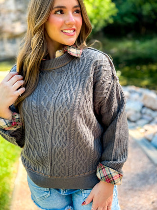 A woman wearing a dark gray cable-knit sweater with a plaid collared shirt underneath, paired with blue jeans, photographed outdoors on a path with greenery in the background.