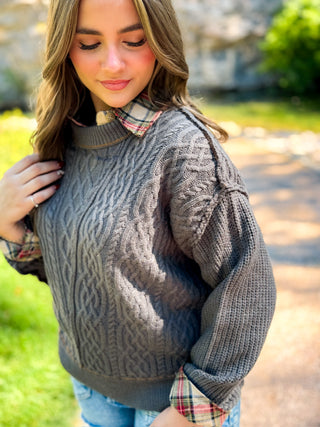 A woman wearing a dark gray cable-knit sweater over a red and green plaid collared shirt, standing outdoors in a sunny garden setting.