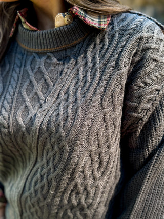 Close-up of a person wearing a textured cable-knit sweater in a warm gray-brown tone, layered over a plaid collared shirt.