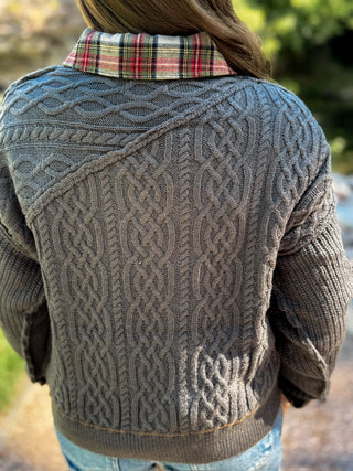 A person viewed from behind wearing a dark gray cable-knit cardigan over a red, green, and black plaid collared shirt, with blue jeans and an outdoor blurred background.