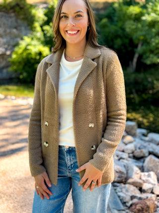A woman smiling outdoors wearing a taupe-brown fuzzy double-breasted coat over a cream top and blue jeans.