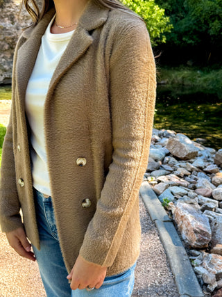 A woman wearing a camel brown fuzzy double-breasted blazer over a white top and blue jeans, standing outdoors next to a stone path and a stream.