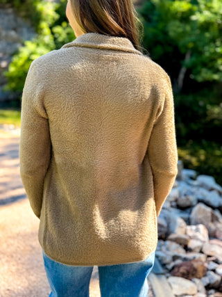 A person viewed from behind wearing a camel-colored, textured fleece or sherpa jacket and blue jeans, standing outdoors on a path with rocks and green foliage in the background.