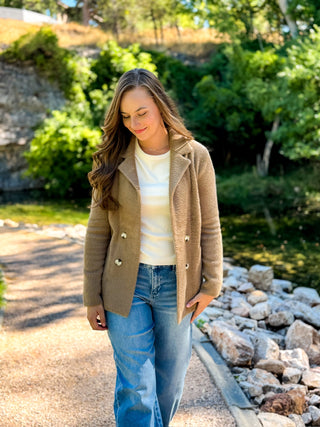 A woman wearing a tan double-breasted knit blazer over a cream striped t-shirt and blue jeans, walking outdoors on a stone path with greenery and rocks in the background.