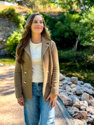 A woman wearing a tan double-breasted knit cardigan over a cream top and blue jeans, standing outdoors on a stone path with greenery and rocks in the background.