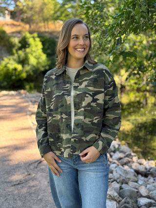 A woman wearing a green and brown camouflage button-up shirt jacket over a light gray t-shirt and blue jeans, standing outdoors on a path with trees and rocks in the background.
