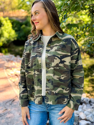A woman wearing a green and brown camouflage button-up shirt jacket over a white t-shirt and blue jeans, standing outdoors in front of a blurred background of trees and rocks.