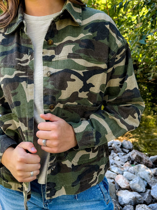 A person wearing a green and black camouflage flannel button-up shirt over a light beige t-shirt and blue jeans, standing outdoors near rocks and green foliage.