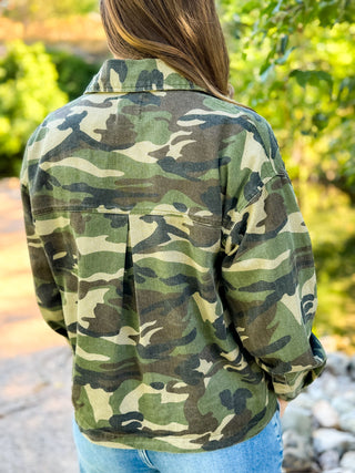 A person viewed from behind wearing a long-sleeve camouflage flannel shirt and light blue jeans, standing outdoors with a blurred green garden background.