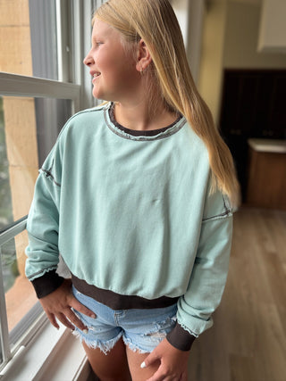 Person wearing a light blue sweatshirt and denim shorts standing by a window.