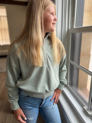Person wearing a green sweater and blue jeans standing by a window.