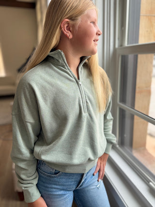 Person wearing a light gray half-zip sweatshirt and blue jeans standing by a window.