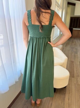 Woman wearing a green dress standing in a room with white curtains and a beige chair.