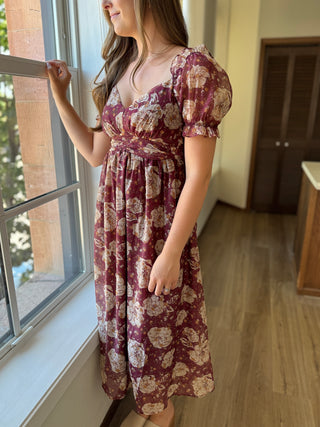 Woman wearing a floral dress standing by a window indoors.