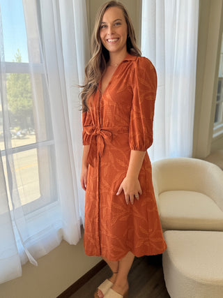 Woman wearing an orange dress standing in a room with white curtains and a beige sofa.