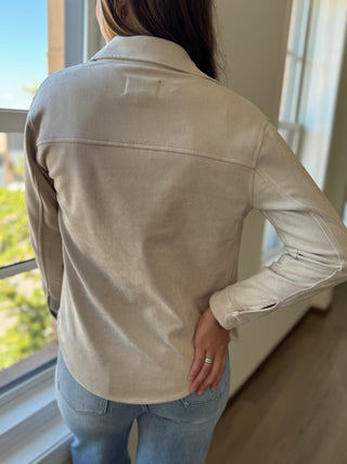 Person wearing a beige jacket standing by a window with a view of greenery outside.