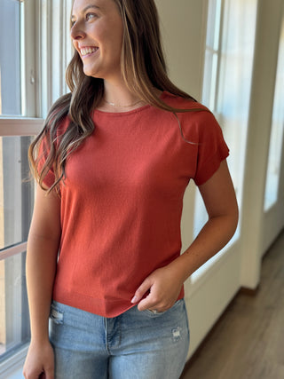 Woman wearing a red shirt and blue jeans standing indoors near a window.