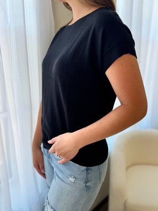Person wearing a black t-shirt and light blue jeans against a white curtain background