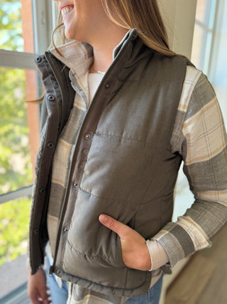 Person wearing a plaid shirt and brown vest with a blurred outdoor background