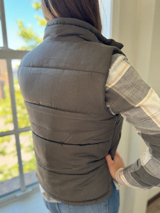 Person wearing a gray puffer vest over a plaid shirt with a blurred outdoor background