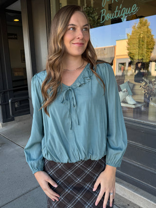 A woman wearing a dusty teal blue long-sleeve blouse with a ruffled tie neckline, standing outdoors in front of a boutique storefront.