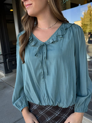 A woman wearing a dusty teal blue long-sleeve blouse with a ruffled V-neck and tie detail, paired with a brown plaid skirt, photographed outdoors in front of a storefront.