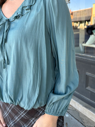 A close-up of a person wearing a dusty teal long-sleeve blouse with a ruffled V-neckline and tie detail, paired with a brown plaid skirt, set against an outdoor storefront background.