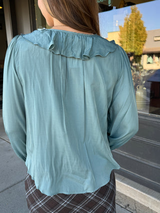 A woman viewed from behind wearing a sage green long-sleeve blouse with a ruffled collar, paired with a brown plaid skirt, standing outdoors near a storefront window.